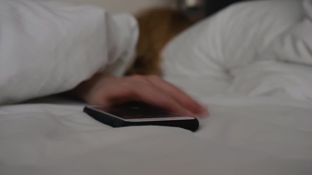Smartphone lying on a white bed and ringing alarm clock. Sleeping tired girl hardly wakes up and tries to turn off the alarm.