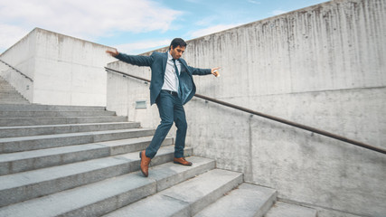 Cheerful and Happy Businessman in a Suit is Holding Coffee and Actively Dancing While Walking Down the Stairs. Scene Shot in an Urban Concrete Park Next to Business Center. Sunny.