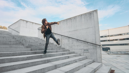 Cheerful and Happy Young Man with Long Hair Actively Dancing While Walking Down the Stairs. He's Wearing a Brown Leather Jacket. Scene Shot in an Urban Concrete Park Next to Business Center. Sunny.