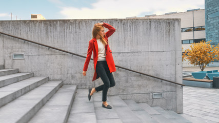 Cheerful and Happy Young Woman Actively Dancing While Walking Down the Stairs. She's Wearing a Long Red Coat. Scene Shot in an Urban Concrete Park Next to Business Center. Day is Bright.