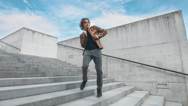 Cheerful and Happy Young Man with Long Hair Actively Dancing While Walking Down the Stairs. He's Wearing a Brown Leather Jacket. Scene Shot in an Urban Concrete Park Next to Business Center. Sunny.