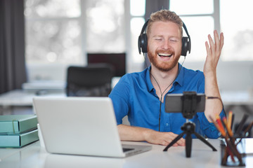 Young Caucasian employee having video call over smart phone while sitting in office. On head are headphones and on desk is laptop and coffee.