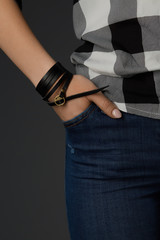 Cropped shot of a female wrist with a multilayer bracelet made of long black leather strip with a bronze rivet and buckle fastener with ornamental holes. The fashion model is dressed in jeans  © RedUmbrella&Donkey