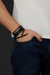 Cropped shot of a male wrist with a multilayer bracelet made of long black leather strip with a bronze rivet and buckle fastener with ornamental holes. The fashion model is dressed in jeans  © RedUmbrella&Donkey