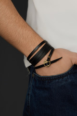 Cropped close-up shot of a male wrist with a multilayer bracelet made of long black leather strip with a bronze rivet and buckle fastener with ornamental holes. The fashion model is dressed in jeans  © RedUmbrella&Donkey