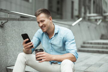young handsome office worker holding smartphone and drinking coffee during lunch break sitting on...