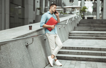 young handsome office worker talking on the phone holding laptop standing on the stairs in the street during lunch break