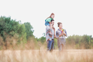 Fototapeta premium Young Caucasian family walking across field with young child on her fathers shoulders with the wife holding a bouquet of flowers
