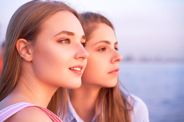 Close up shot of two attractive young women, sitting on sea shore on beautiful summer evening, enjoying amazing colors of sunset, looking in the distance with pleasant smiles, hope and delight.