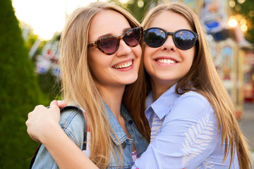 A close shot of two attractive long haired young women, wearing stylish sun glasses, happily looking at camera with gorgeous smiles, hugging each other. Long awaited meeting of two close friends.