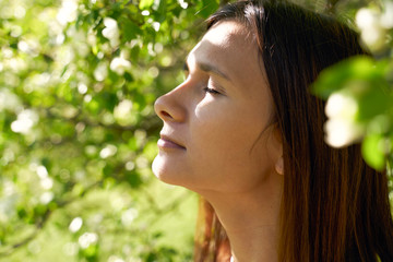 Young cute dark haired girl, illuminated by sunshine, standing in apple trees with her eyes closed,...