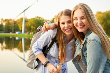 Two pretty student girls, dressed casually, enjoy walking in city amusement park after their college exam, eat ice-cream, pose cheerfully at camera with broad gorgeous smiles, being in joyful mood.