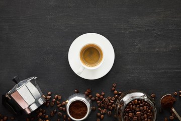 Top view of cup of espresso on saucer near geyser coffee maker, glass jar and spoon on dark wooden surface with coffee beans