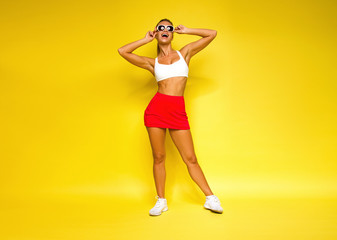 young sports happy sexy blonde girl in red skirt and white top is posing like model excited and looking up with sun glasses in her hands on yellow wall background, sport lifestyle concept, free space