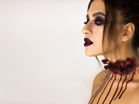 Close-up portrait of young stylish sexy brunette girl with dark bloody makeup with drops which is standing and looking straight on white wall background, halloween art concept, free space on left side