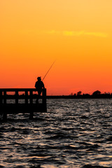 Fototapeta na wymiar Silhouette of a fisherman looking off into the distance across the ocean, as the sunset lights up the sky with an orange glow. Jones Beach State Park Fishing Piers, Long Island New York. 