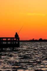 Silhouette of a fisherman looking off into the distance across the ocean, as the sunset lights up the sky with an orange glow. Jones Beach State Park Fishing Piers, Long Island New York. 