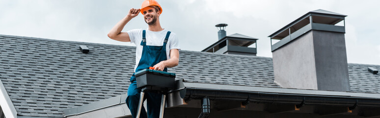 panoramic shot of happy repairman sitting on roof and holding toolbox