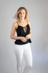 Full length studio portrait photo of a cute young blonde woman girl in a black blouse and white pants on a white background. He stands right in front of the camera, explains with emotion.