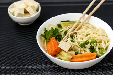 Traditional asian soup with tofu cheese, noodles, carrots and zucchini on dark background. This dish usually contains bouillon and vegetables