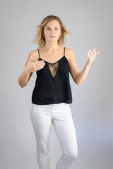 Full length studio portrait photo of a cute young blonde woman girl in a black blouse and white pants on a white background. He stands right in front of the camera, explains with emotion.