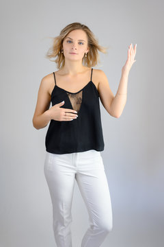 Full length studio portrait photo of a cute young blonde woman girl in a black blouse and white pants on a white background. He stands right in front of the camera, explains with emotion.