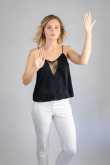 Fototapeta premium Full length studio portrait photo of a cute young blonde woman girl in a black blouse and white pants on a white background. He stands right in front of the camera, explains with emotion.