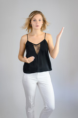 Full length studio portrait photo of a cute young blonde woman girl in a black blouse and white pants on a white background. He stands right in front of the camera, explains with emotion.