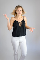 Full length studio portrait photo of a cute young blonde woman girl in a black blouse and white pants on a white background. He stands right in front of the camera, explains with emotion.