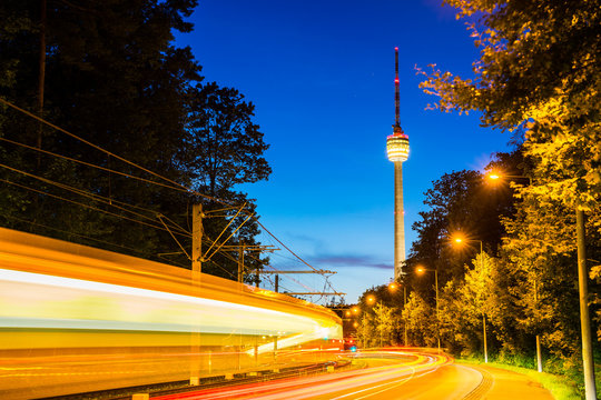 Germany, Stuttgart television tower and tramway illuminated by night at road with traffic and starry sky in summer surrounded by forest nature landscape