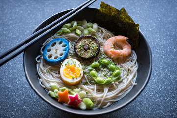 Bowl of Japanese buckwheat ramen noodle soup topped with colorful circles of blue lotus root, shiitake mushroom, shrimp, boiled egg, and edamame beans