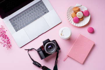 Overhead image of workplace with laptop, coffee, camera and macarons on pink background. Top view, copy space. Creativity concept