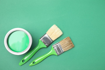 Open can with paint and brushes on green background, flat lay. Space for text