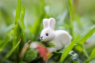 A white rabbit toy sitting on the ground in the grass, leaves and flowers