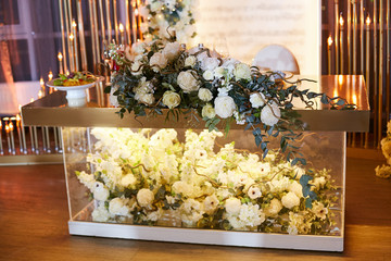 A luxurious wedding table of the newlyweds made of glass with a gilded tabletop, decorated with bouquets of beautiful white flowers and greenery. Garland with a gentle light in the background. 