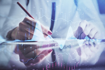 A woman hands writing information about stock market in notepad. Forex chart holograms in front. Concept of research.