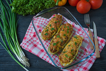 Stuffed zucchini with minced meat and grated cheese in a glass baking sheet. Dark wooden background.