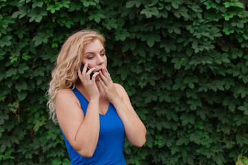 Beautiful young girl in a blue dress talking on the phone. Against the background of wild grapes, summer day. Copy space.