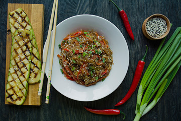 Asian cuisine. Cellophane noodles decorated with vegetables, greens. Funchoza. Proper nutrition. Healthy food. View from above. Dark wooden background.