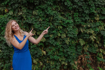 Young beautiful girl on a background of a wall of wild grapes, points fingers to the side. Copy space.