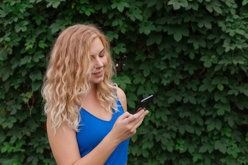Beautiful young girl in a blue dress typing a message on the phone. Against the background of wild grapes, summer day. Copy space.