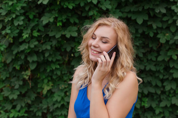 Beautiful young girl in a blue dress talking on the phone. Against the background of wild grapes, summer day. Copy space.
