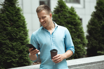 young handsome smiling man drinks coffee on the go standing near office business center checking...