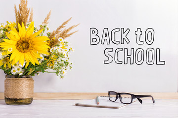 The inscription marker on a white board, Back to School. A table with a bouquet of flowers, glasses and attributes for writing.