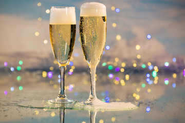 Yellow and bubbly champagne served in two glass cups against a background illuminated by many coloured lights. Concept of party and celebration.