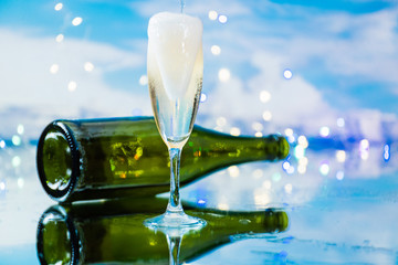 Sparkling champagne served in a glass cup in front of a green bottle of champagne on a background illuminated by many small coloured lights. Concept of party and celebration.