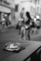 black and white, an ashtray with cigarette butts on a restaurant table on an evening city street, a vase and a menu, people in the background. smoking concept, harmful to health, vertical photo