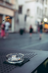 vertical photo, an ashtray with cigarette butts on a restaurant table on an evening city street, a vase and a menu, people in the background. smoking concept, harmful to health