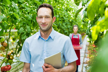Smiling confident supervisor standing while holding digital tablet in greenhouse