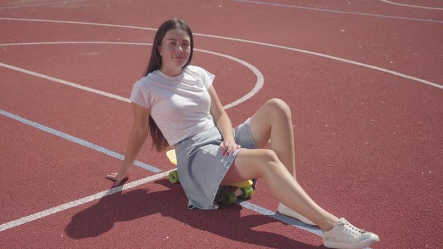 Attractive young woman in short skirt sitting on the skateboard and looking at the camera on the basketball court. Concept of sport, power, competition, active lifestyle